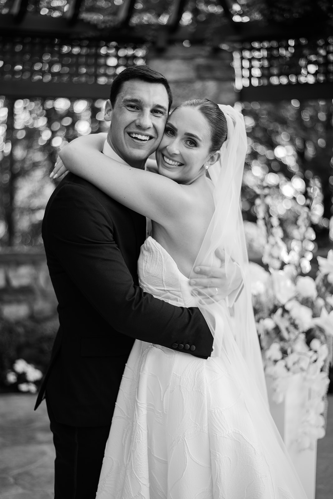 Black and white film portrait of the newlyweds hugging and smiling joyfully at the camera.